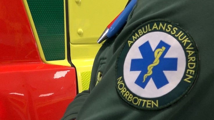 Bilden visar en person som st&aring;r framf&ouml;r en ambulans med ambulanssjukv&aring;rds logotypen